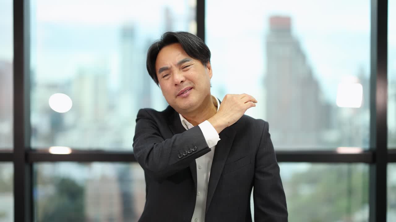 Businessman in suit massages neck and shoulder, expressing discomfort, in bright modern office setting