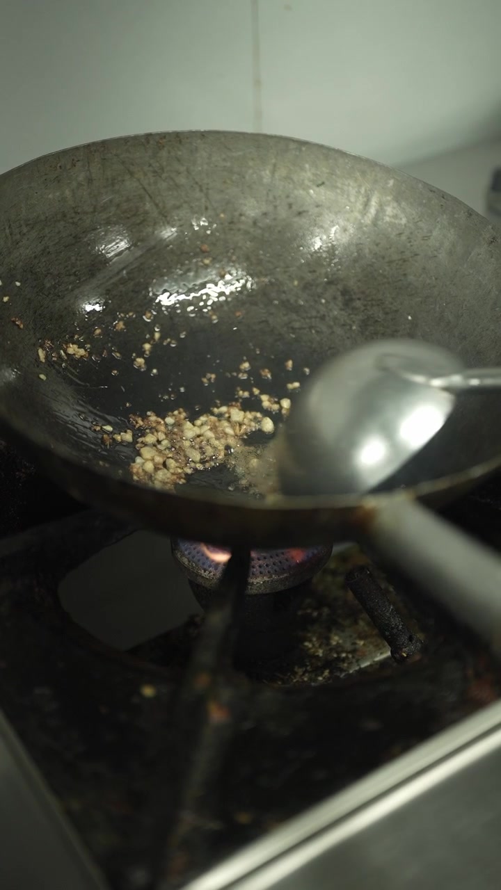 Riprese verticali ravvicinate di aglio tritato leggermente saltato, distribuito su un wok di ghisa caldo con burro fuso, utilizzando un mestolo da cucina su una fiamma a gas
