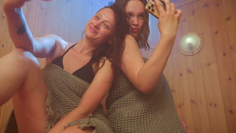 two women wrapped in towels sit on wooden sauna bench surrounded by steam haze, one holding smartphone for selfie as they make playful faces under warm ambient light