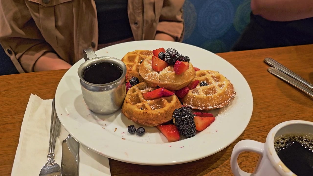 Primer plano de mini gofres con fresas, moras, arándanos, azúcar glas, taza de sirope y café en una mesa de restaurante para videos de brunch, desayuno, cafetería y postres.