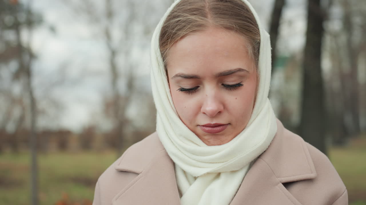 Close-up wanita Kaukasia bersyal, senyuman tipis muncul, taman musim gugur dengan pohon-pohon gundul, palet warna kalem, mantel krem dan syal lembut, tatapan merenung ke bawah, angin sejuk, cahaya alami yang lembut.