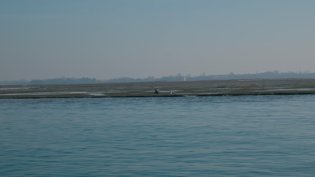 베네치아에 있는 배에서 내려다보는 평온한 바물과 은 파란 하늘 아래 멀리 떨어진 낮은 섬을 포착하는 조용한 풍경.