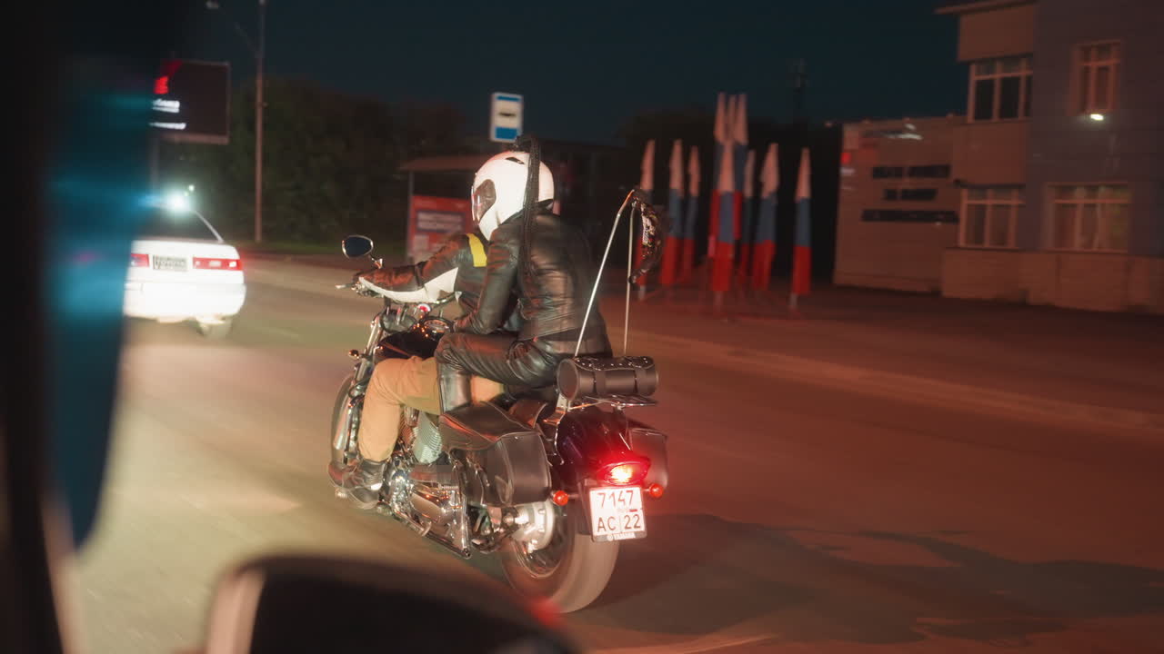 Couple wearing helmets and leather jackets riding motorcycle on busy urban street at night, illuminated by headlights and city lights, passing cars and buildings during road trip