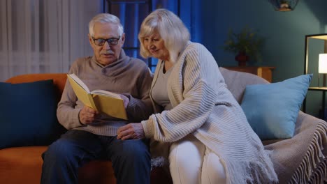 senior grandparents couple relaxing, reading book, talking enjoying leisure hobbies at evening home