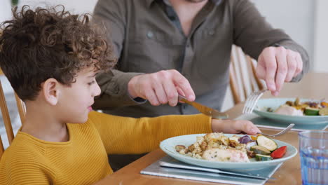 Alleinerziehender-Vater-Hilft-Seinem-Sohn-Beim-Schneiden-Von-Essen,-Während-Sie-Zu-Hause-Am-Tisch-Sitzen-Und-Essen