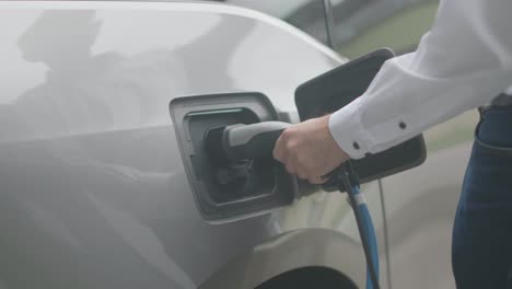 Close-up-of-a-modern-electric-car-charger-being-attached-to-a-silver-electric-car-by-a-male-person-wearing-a-white-shirt