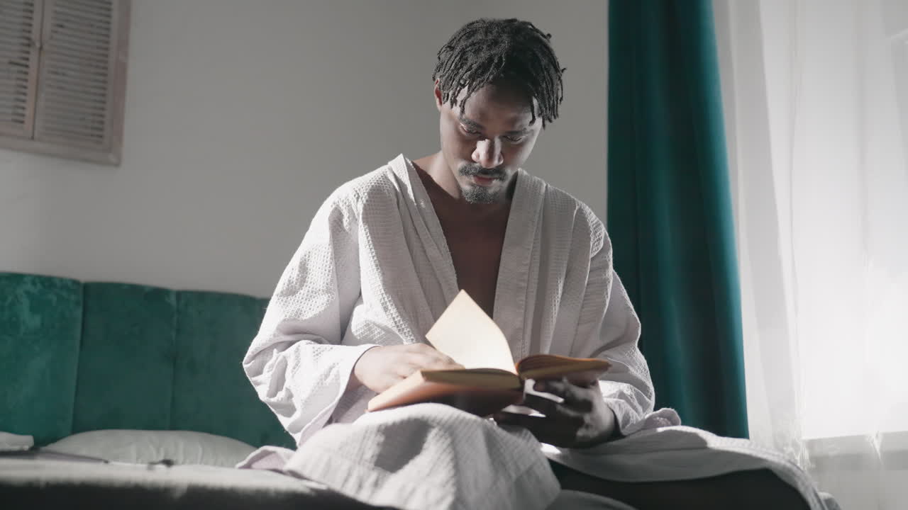 Guy in white bathrobe sitting on bed, reading book with focus. Morning sunlight fills room, creating soft, tranquil atmosphere. Cozy indoor setting with peaceful mood and relaxed vibe