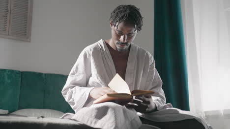 guy in white bathrobe sitting on bed, reading book with focus. morning sunlight fills room, creating soft, tranquil atmosphere. cozy indoor setting with peaceful mood and relaxed vibe