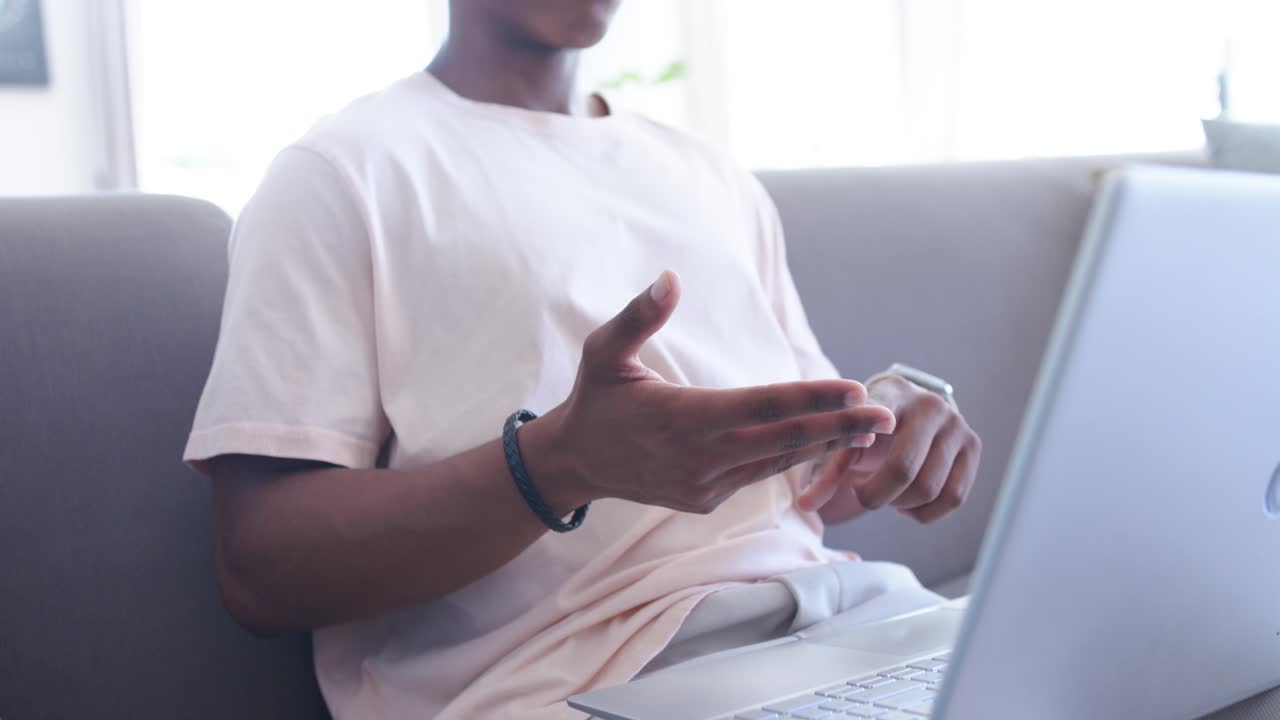 African adult man sitting on grey sofa, gesturing at laptop during online call, checking smartwatch