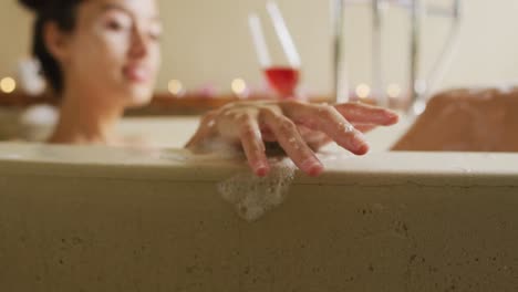 Hands-of-happy-biracial-woman-with-vitiligo-lying-in-bath-with-foam-and-a-glass-of-wine