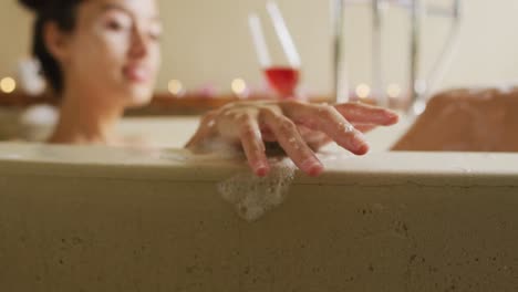 hands of happy biracial woman with vitiligo lying in bath with foam and a glass of wine