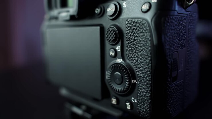 Close-up of mirrorless interchangeable-lens camera body with flip-out LCD screen and control dials in moody studio lighting, shallow depth of field