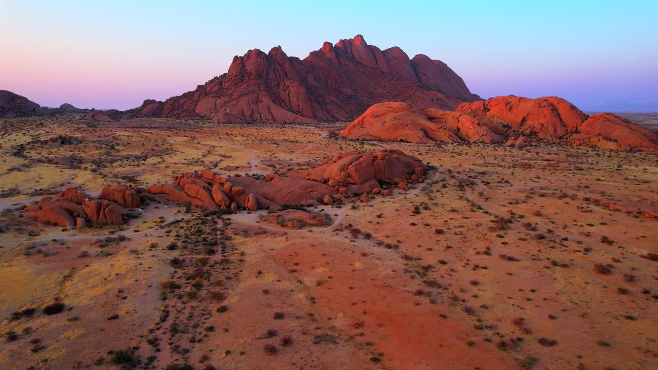 Drone pull back over reddish granite boulders reveals towering inselbergs of Spitzkoppe under sunset sky in Namibia