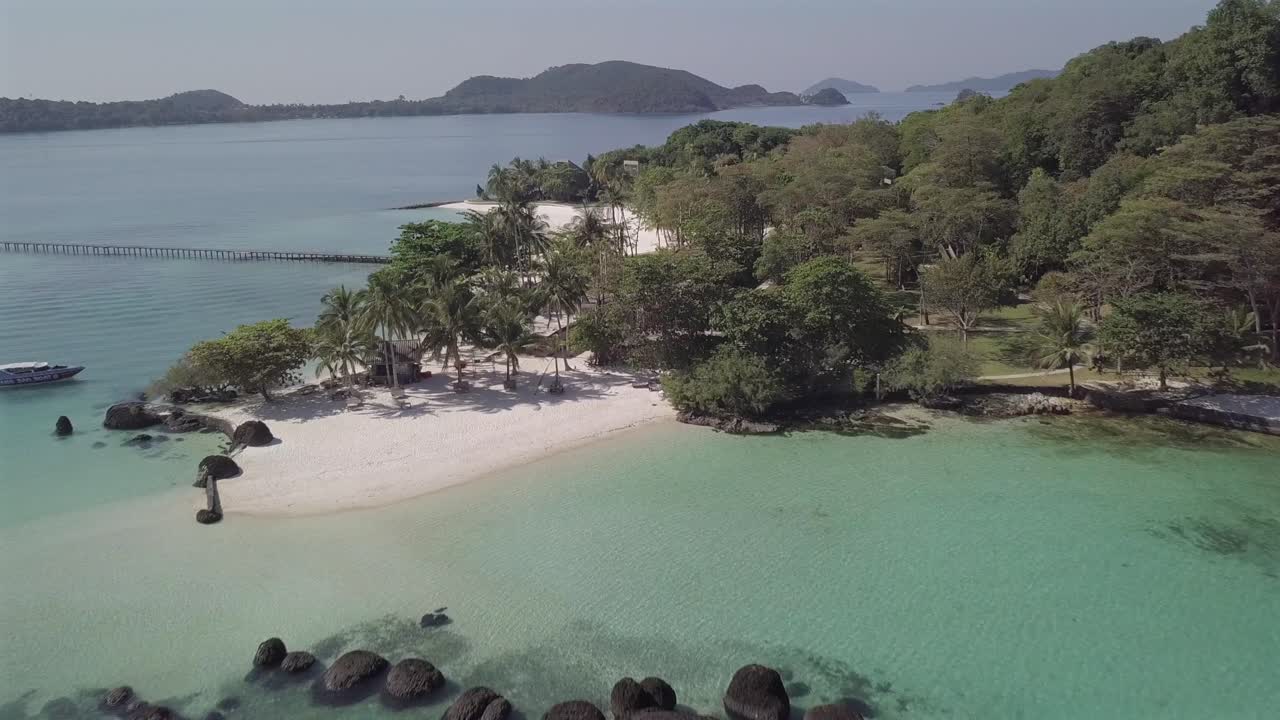 Aerial view forward over black stones in turquoise waters towards tropical white sand beach and palm trees of paradise island Koh Kham,Thailand.