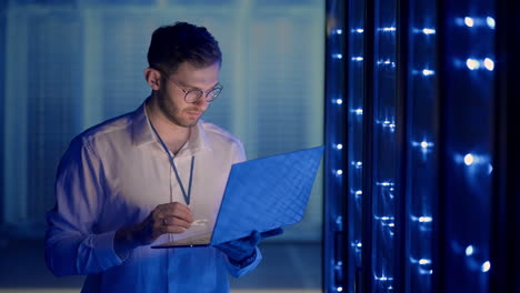 Male-network-engineer-doing-a-system-check-standing-in-the-server-room-with-his-laptop.-At-data-center-men-server-specialists-inspecting-working-system-and-hardware-of-rack-server-computer-cabinets