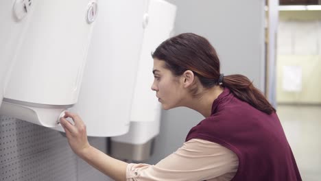 Young-girl-looking-for-water-heater-turns-the-button-on-white-boiler-and-examines-it-in-hardware-store.-Costumer-looking-for-house-appliance.