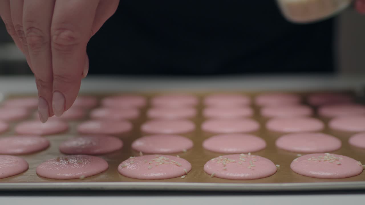 el chef de confitería salpica con semillas de sésamo las galletas de macarrón sin cocinar que se encuentran en la hoja de hornear. cámara lenta.