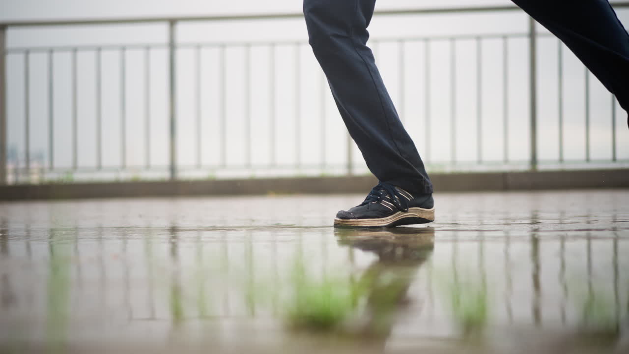 Tejado mojado con zapatillas chapoteando, persona solitaria caminando por la barandilla metálica bajo una suave llovizna, charcos que reflejan el cielo nublado, movimiento cinematográfico lento que captura cada paso, ambiente sutilmente melancólico