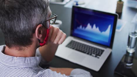 Kaukasischer-Mann-Mit-Gesichtsmaske-Und-Laptop,-Während-Er-Auf-Seinem-Schreibtisch-Im-Modernen-Büro-Sitzt