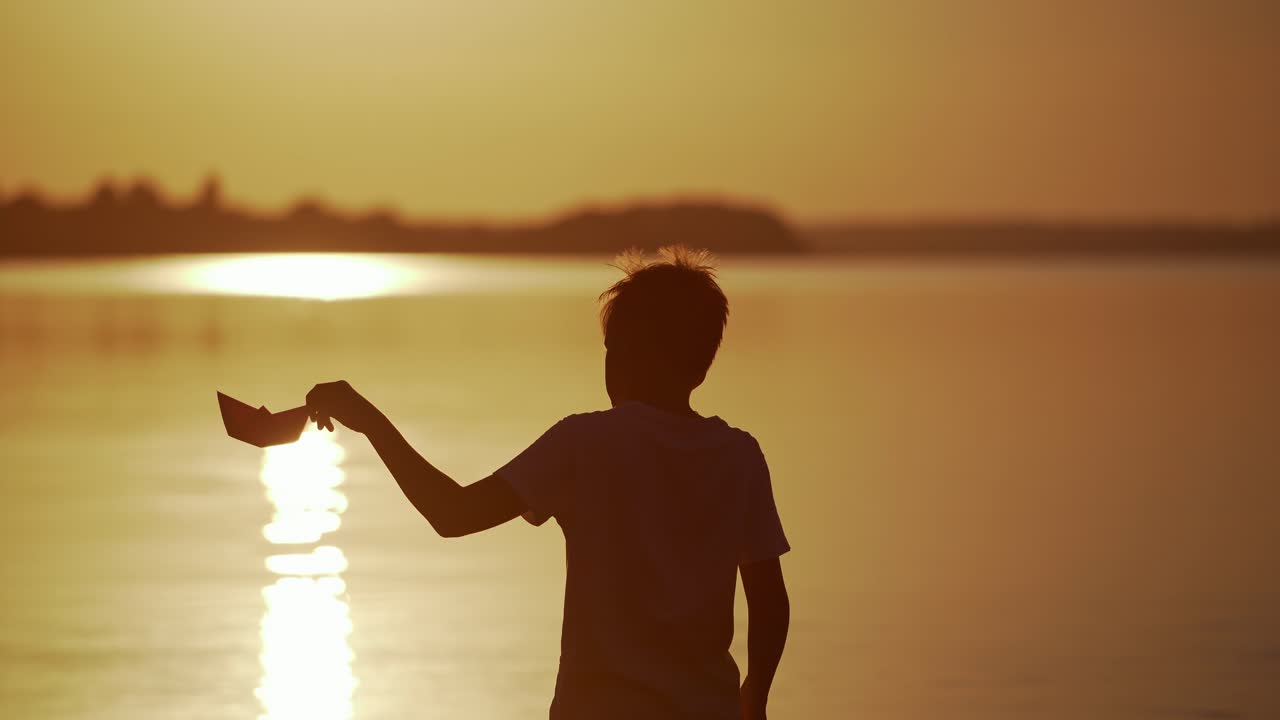 Backside view of a boy at sunset. Silhouette of the boy playing with origami boat on evening water background outdoors. Dreamy childhood.