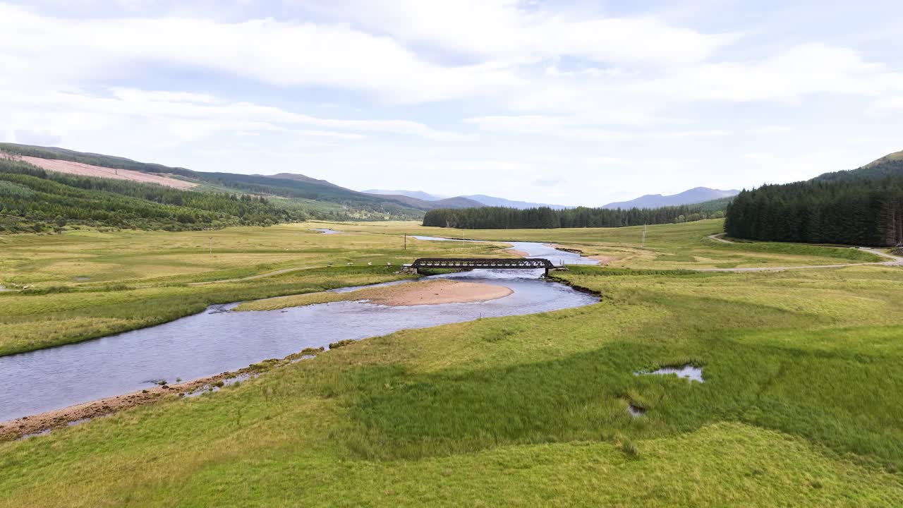 Drone glides above tranquil river, bridge, and lush green valley under soft daylight in Scotland