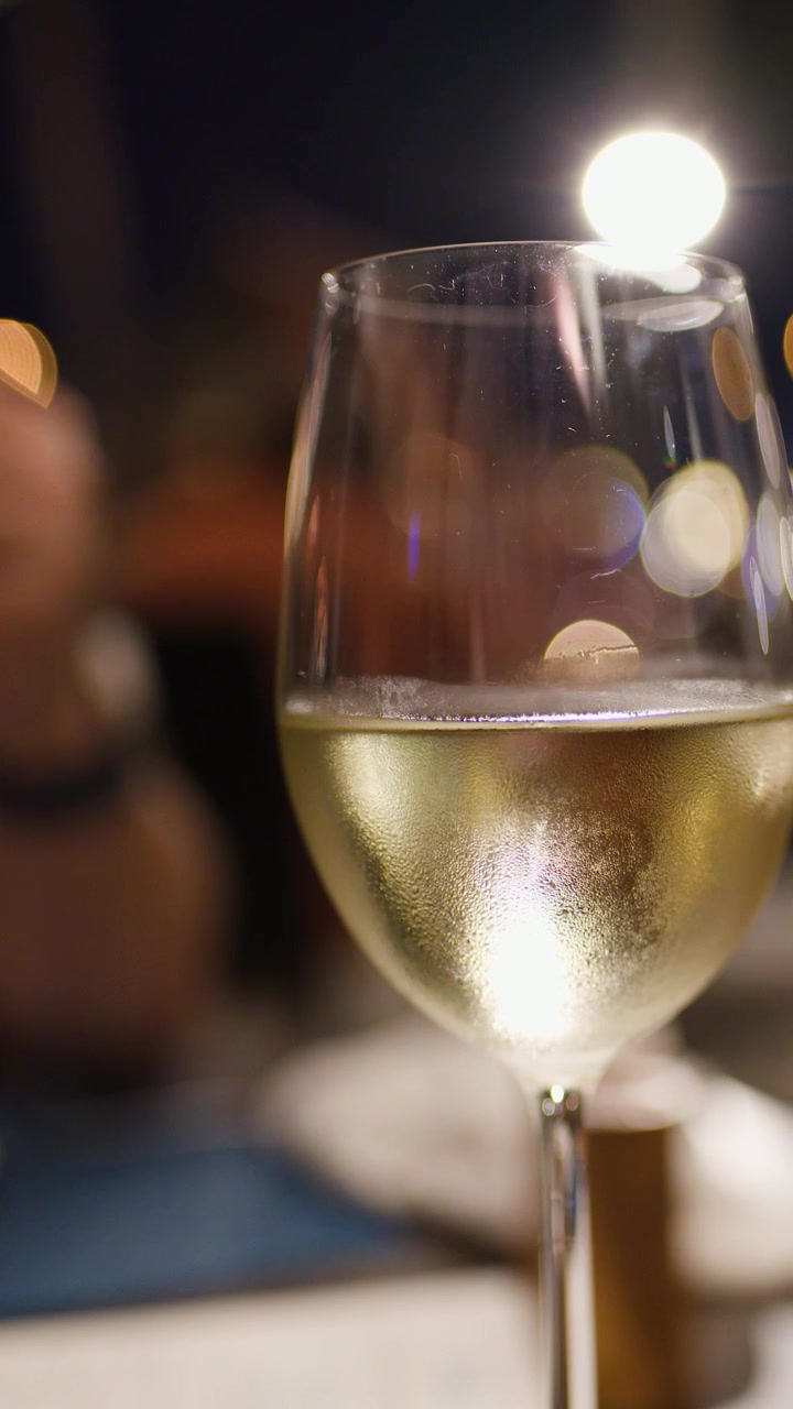 Primo piano di un bicchiere di vino bianco ghiacciato, luci bokeh, cena serale, atmosfera elegante del ristorante.
