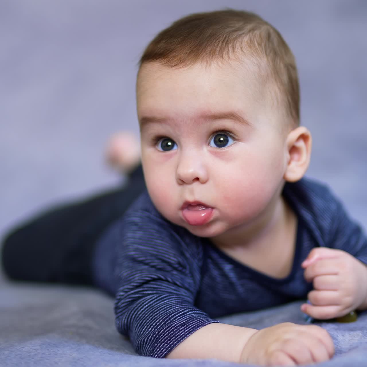 Lovely baby boy lies on belly looking up with surprise. Cute kid starts to move his limbs actively. Close up. Blurred grey backdrop