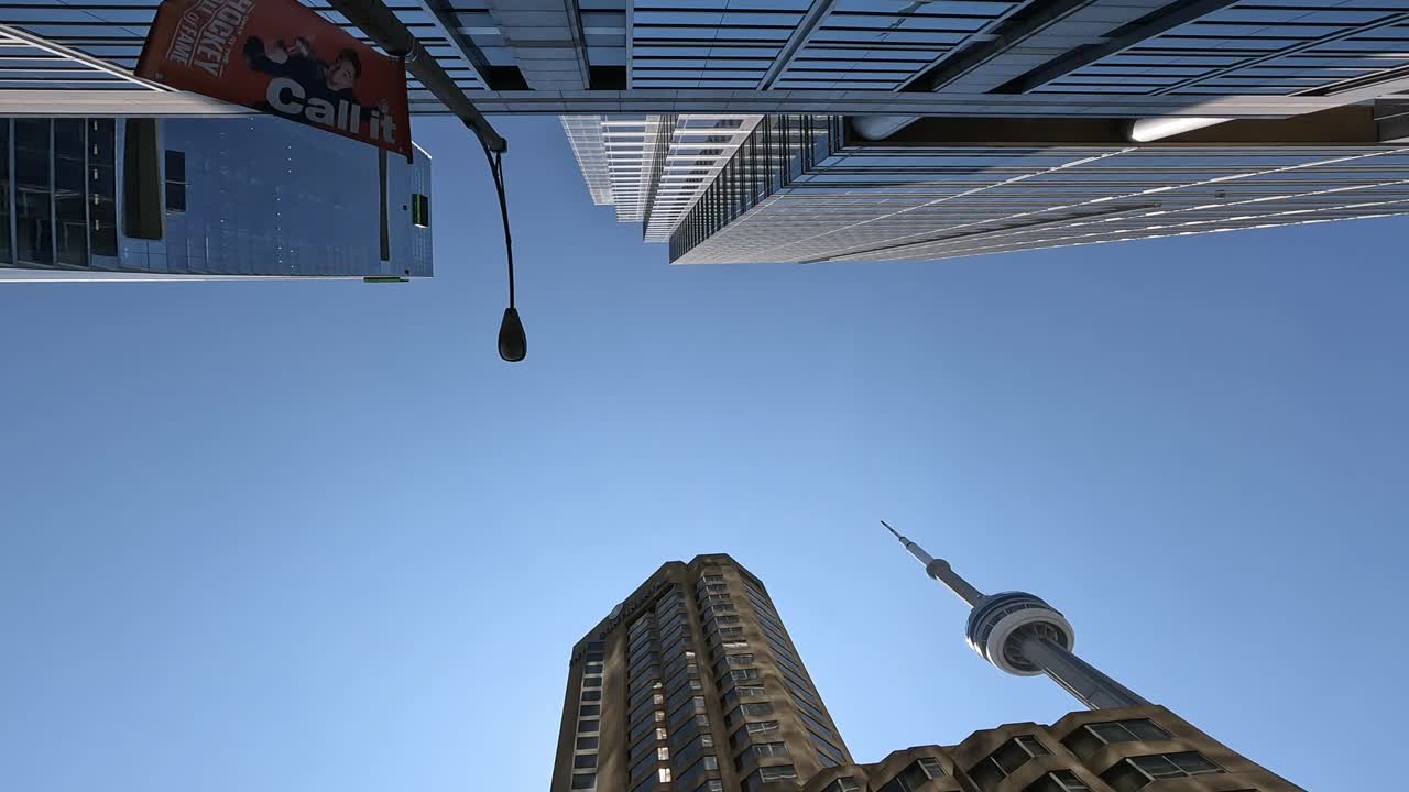 Dramatic upward view of the CN Tower surrounded by modern skyscrapers in downtown Toronto set against a bright blue sky. The architectural framing emphasizes urban grandeur. Canada