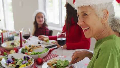 Glückliche-Kaukasische-ältere-Frau-Mit-Weihnachtsmütze,-Die-Während-Des-Weihnachtsessens-In-Die-Kamera-Blickt