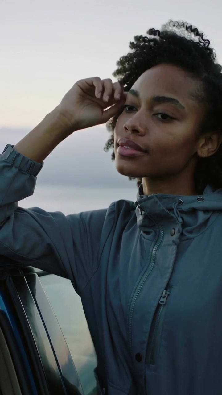 Vertical video: Leaning woman resting chin, fixing hair watching sun at coast hooded jacket car sea