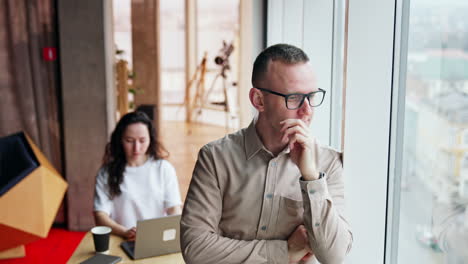 millennials working in the modern office. pensive man stands at the window with his arms crossed on chest. woman sits at desk working at laptop in backdrop.