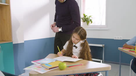 chica concentrada escribiendo en un cuaderno durante la clase de inglés en la escuela mientras el profesor se acerca y revisa su trabajo 1