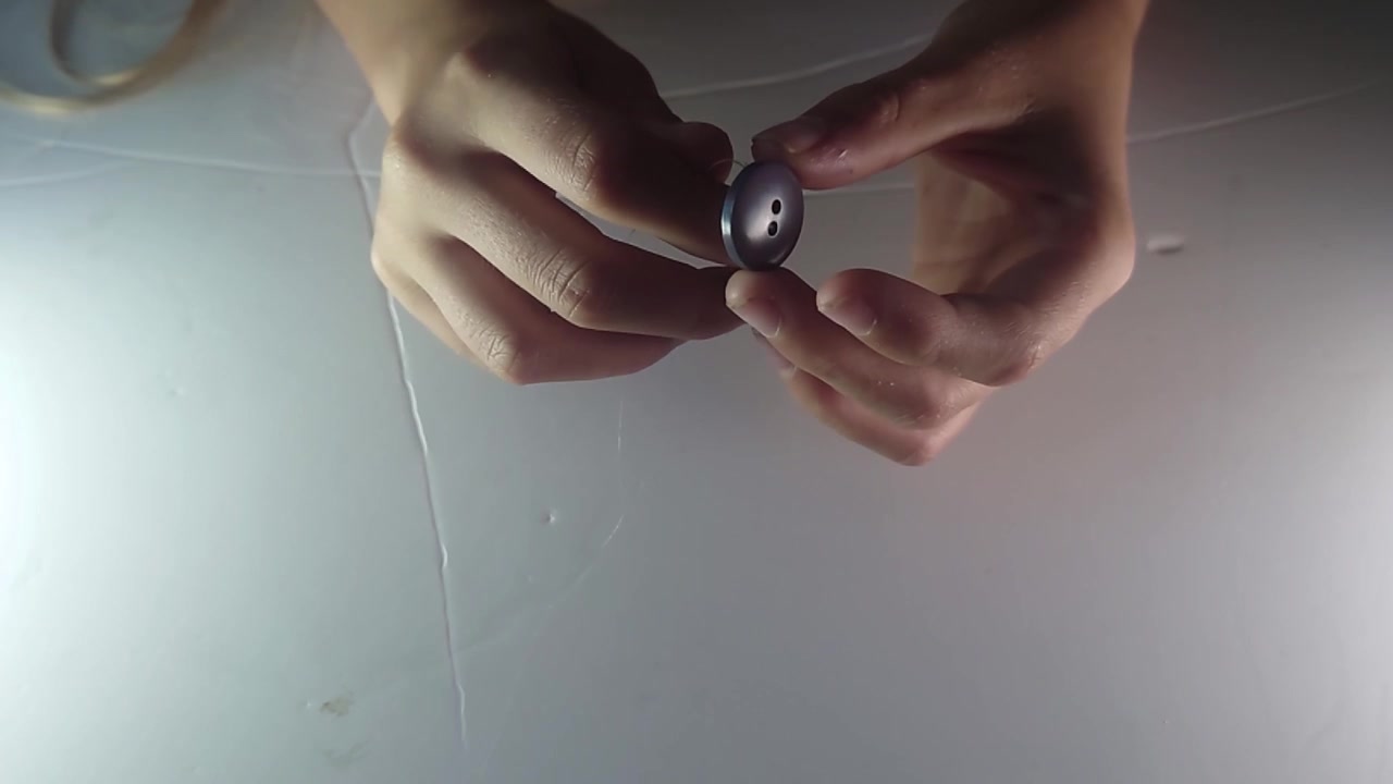 A top-view close-up shot shows human hands carefully threading a thin string through the holes of a dark button to construct a traditional vintage whirlygig spinner toy on a bright white surface.