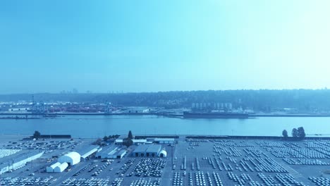 automotive-industry-import-export-lot-next-to-loading-port-for-trade-freighter-dock-at-commercial-area-with-rows-of-cars-lined-up-and-plastic-wrapped-on-a-sunny-smoggy-summer-day-aerial-4k-top-view