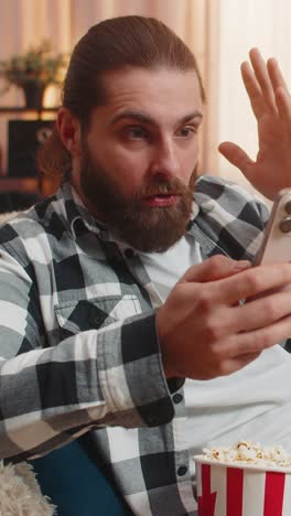 young man on sofa with popcorn smartphone scroll sudden wow reaction shock moment home surprise