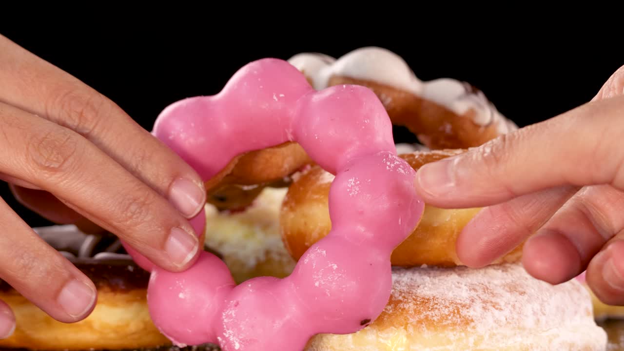 Two hands simultaneously grab and pull apart a pink glazed donut above assorted donuts on a black background, with bright, even lighting and minimal camera movement