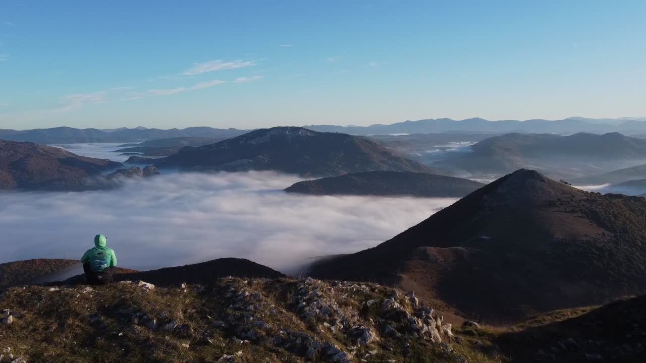 vuelo de drones sobre una persona sentada en la cima de una montaña al amanecer, viendo un mar de nubes a la hora dorada