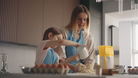 Madre-E-Hija-Están-Cocinando-En-La-Cocina-De-Casa,-La-Mujer-Está-Amasando-Masa-Y-La-Niña-Está-Vertiendo-Agua