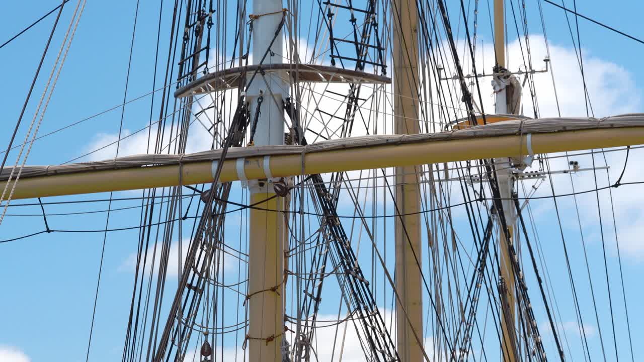 Tall ship rigging gently swaying, backlit against a blue sky with puffy clouds, showing three masts and crow's nest