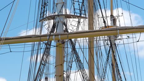 tall ship rigging gently swaying, backlit against a blue sky with puffy clouds, showing three masts and crow's nest