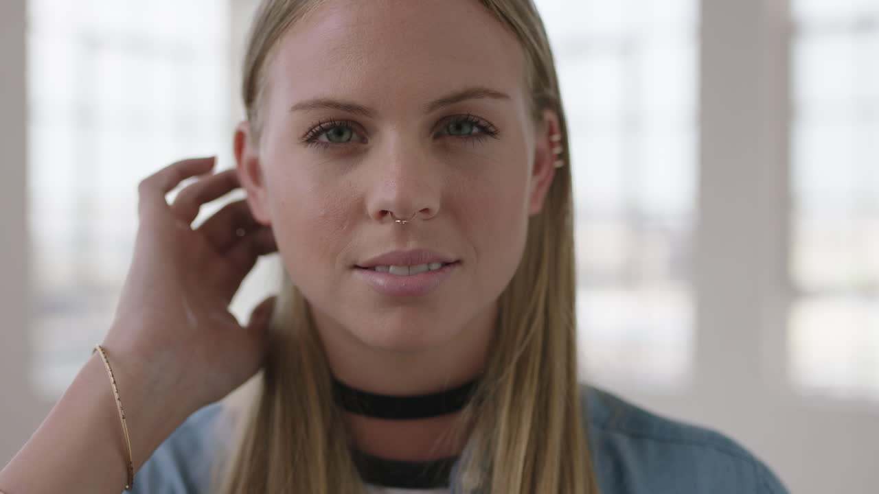close up portrait of beautiful young blonde woman looking serious pensive at camera running hand through hair in apartment windows background