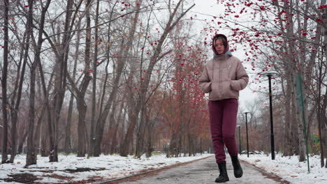 woman in warm winter outfit walking with hands in pockets on an empty snowy street lined with bare trees adorned with red berries, footprints visible on frozen pathway