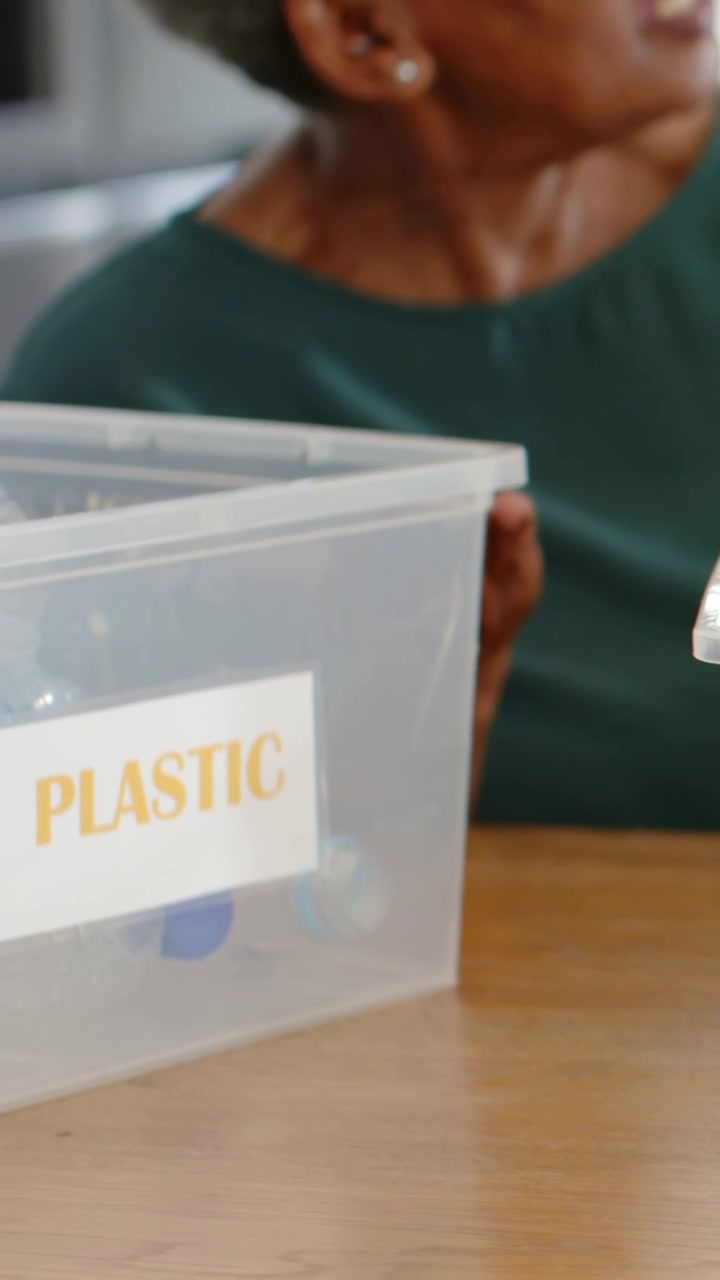 Vertical video: Reaching woman in green lifting plastic bin for recycling beside glass bin at home