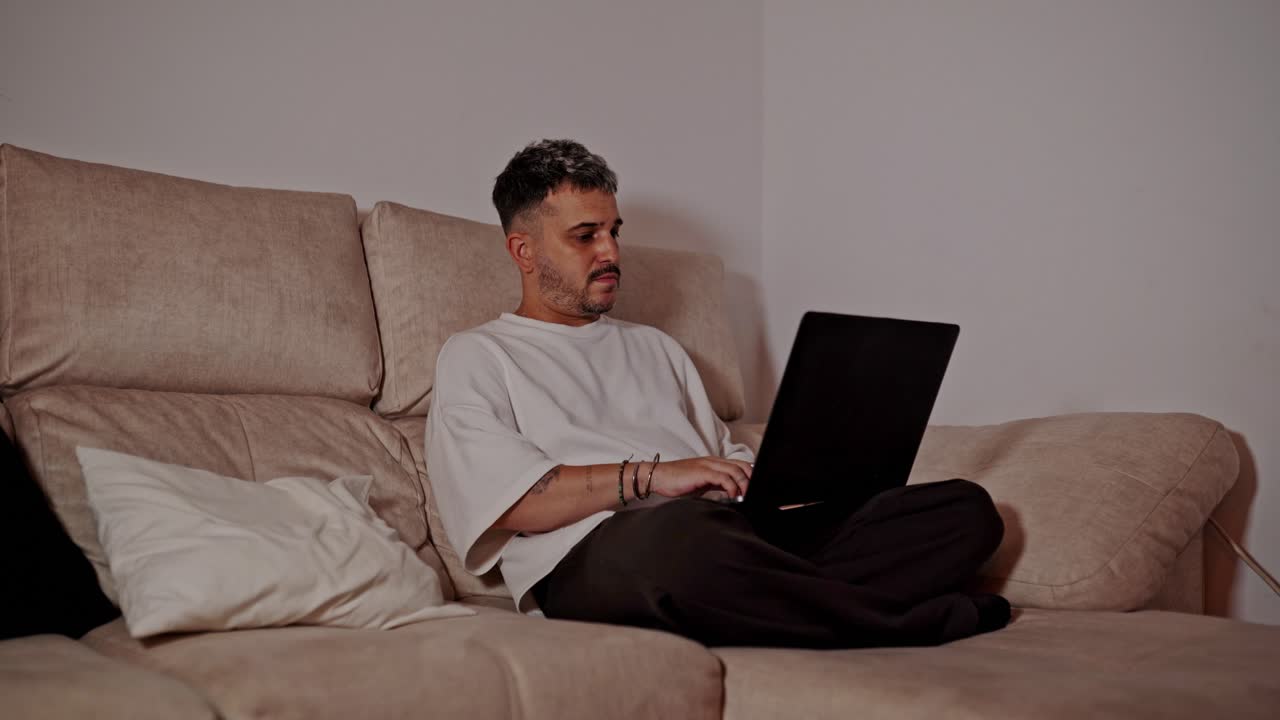Young man sitting cross-legged on couch looking thoughtful while using laptop at home