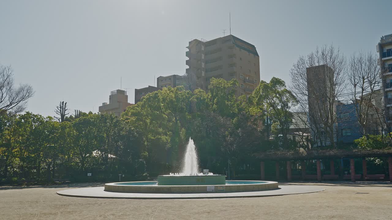 Ampia inquadratura della fontana d'acqua centrale che spruzza nel Parco Gyosen, circondata da alberi verdi ed edifici cittadini sotto un cielo limpido e soleggiato