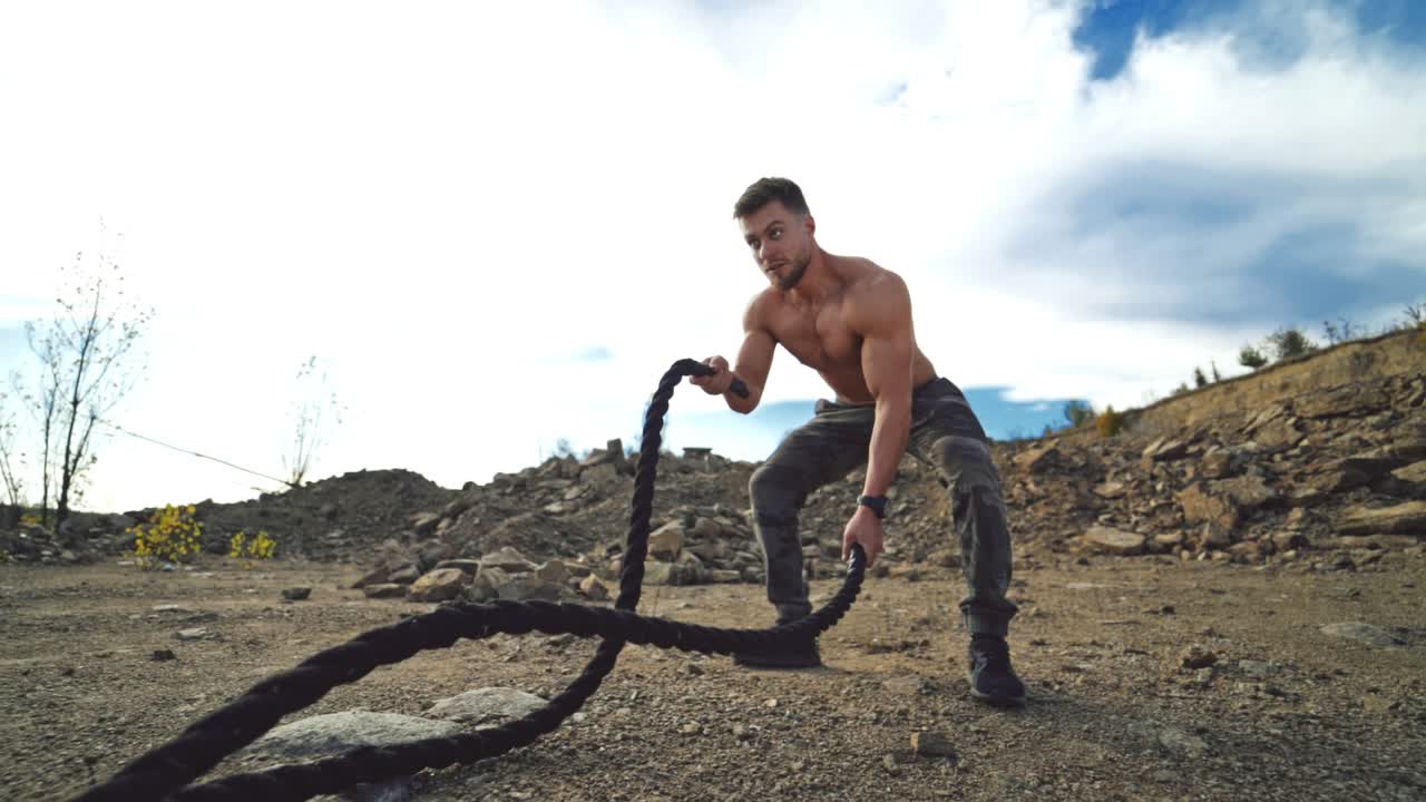 Muscular athlete doing waves with battle ropes in nature. Professional sportsman exercises with cables outdoors. Healthy lifestyle.