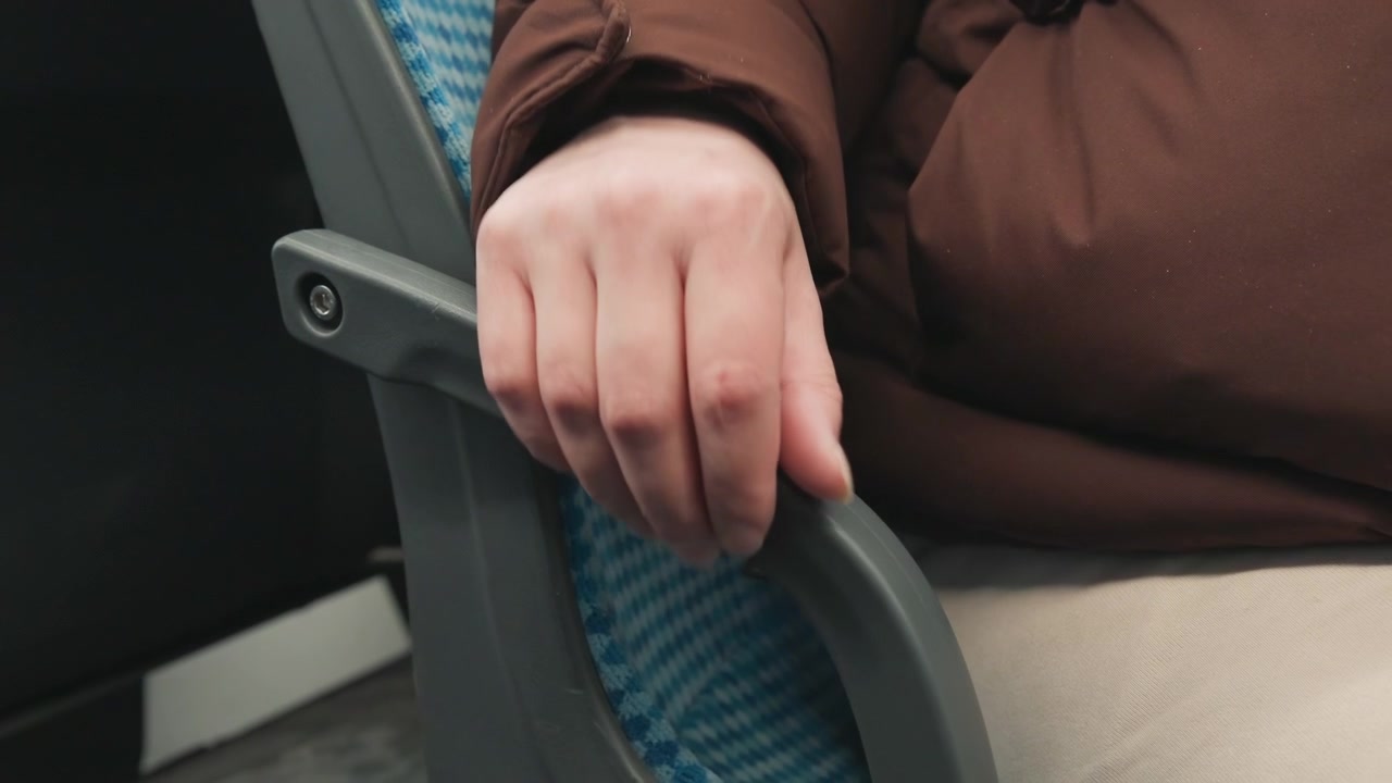Vista lateral cerrada de un reposabrazos de plástico curvado y tapicería azul estampada dentro del interior de un autobús o tren, con la ropa de un pasajero parcialmente visible y un ambiente de descanso tranquilo que sugiere la comodidad del viaje diario.