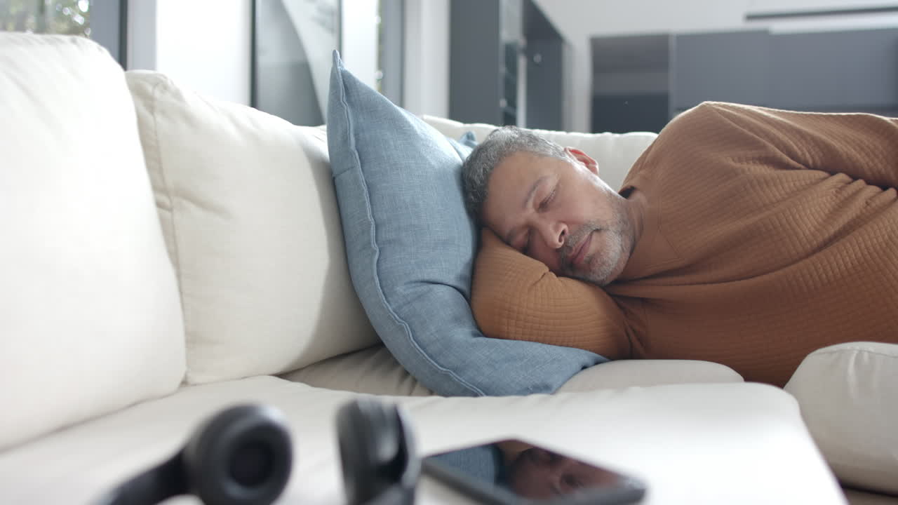 Træt ældre mand af blandet etnicitet ligger og sover på sofaen i solrig stue, kopiplads, slowmotion