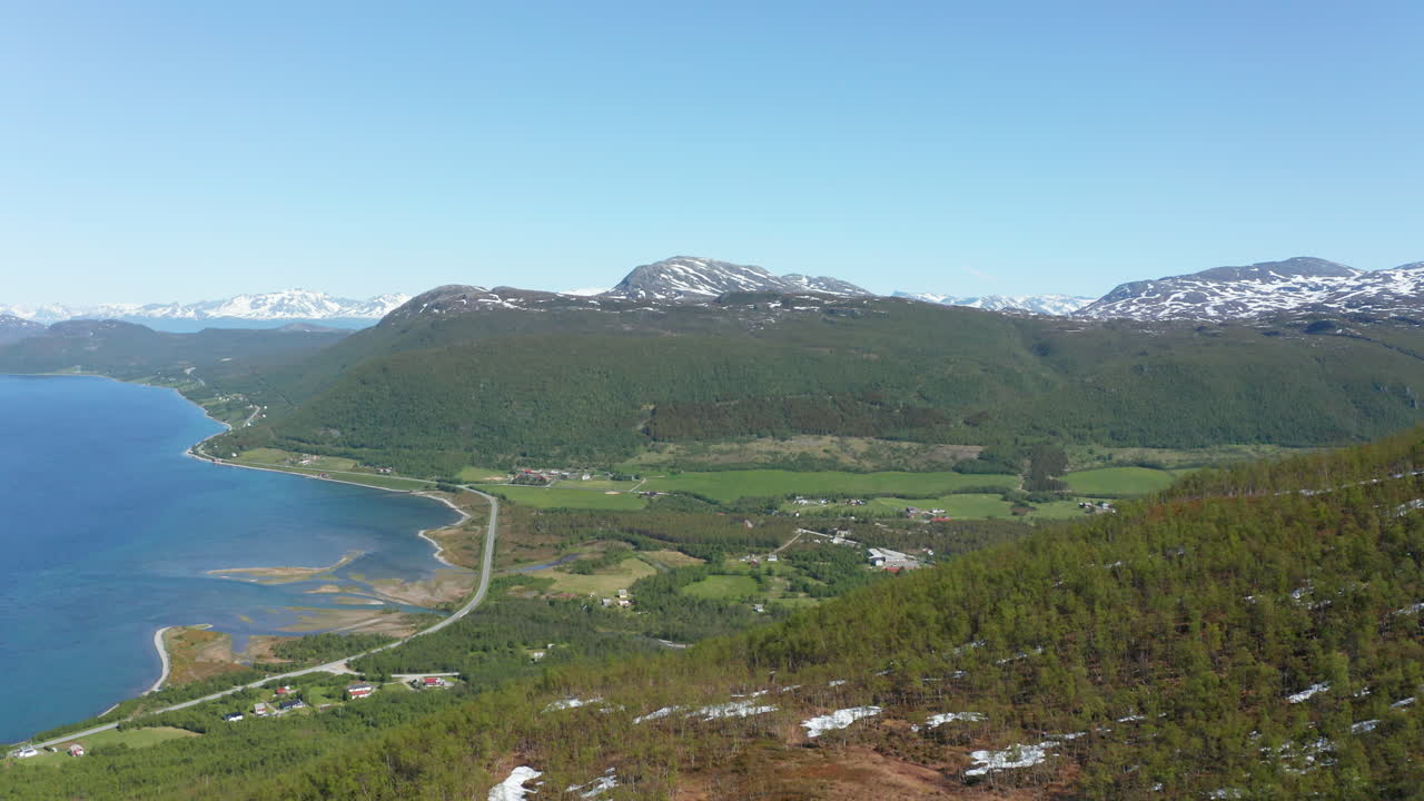 vista aérea en lo alto de una montaña, con vistas a un pueblo y una carretera costera y la costa del océano ártico, soleado, día de verano, en rotsund, troms, nordland, noruega - carro, tiro de drone