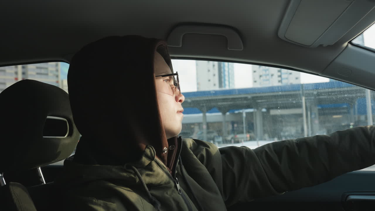 young man in hoodie and glasses sits in parked car side view hand resting on steering wheel dancing gently to lively music through tinted windshield urban winter backdrop sunlight relaxed vibe